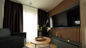 A modern living room with a dark sofa, a round coffee table, a TV mounted on a wooden panel wall, and a window with curtains. - Airport Apartments Zürich (Opfikon)