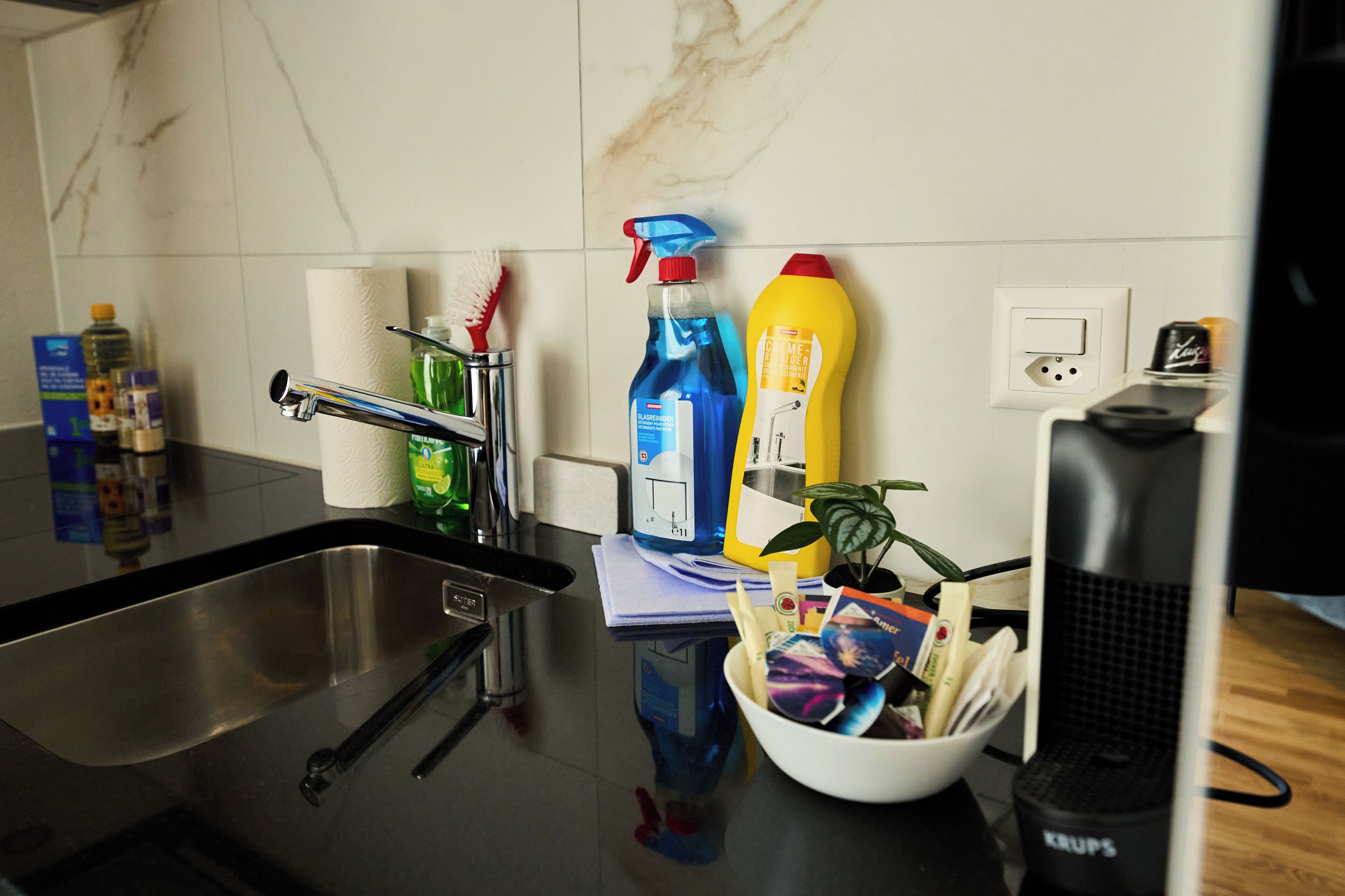 A kitchen with a stainless steel sink, a white countertop, and various cleaning supplies.