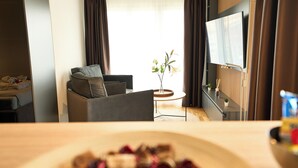 A hotel room with a sofa, a small table with a vase, a flat-screen TV, and a large window with curtains. - Airport Apartments Zürich (Opfikon)