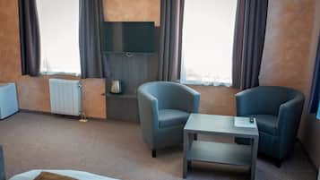 Habitación de hotel con dos sillones, una mesita, televisor montado en la pared, radiador y ventana con cortinas.