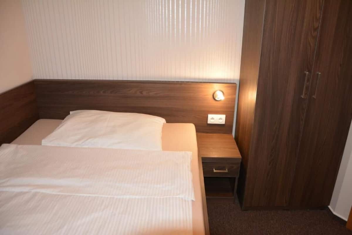 Une chambre d’hôtel avec une tête de lit en bois, un lit simple avec du linge de lit blanc, une table de chevet avec une lampe et une armoire.