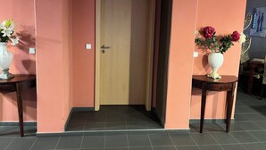 Un pasillo con una puerta de madera, un jarrón blanco con flores sobre una mesa, y un jarrón parecido en la pared opuesta.