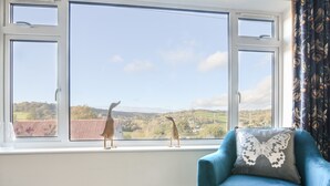 Ein Zimmer mit einem großen Fenster, einem blauen Sessel und zwei hölzernen Tiermotive auf der Fensterbank.