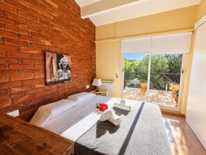 A hotel room with a brick wall, a bed with white linens, a small table with a towel and a flower, and a sliding glass door leading to an outdoor area with plants. - Stunning Villa in Platja d'Aro: 100m from Beach, Terrace, A/C, Family-Friendly (Platja d'Aro)