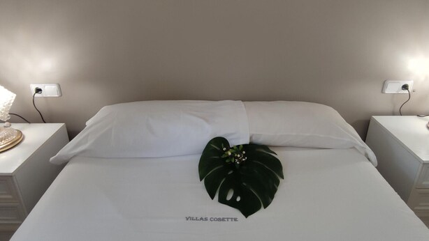A neatly made bed with white linens and a decorative leaf on the center. There are two bedside tables with lamps and a wall socket on each. - Cozy Apartment with Terrace Near Beach and Shops in Costa Brava (Platja d'Aro)