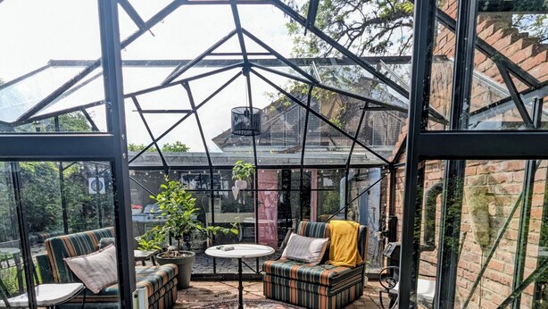 Una serra con tetto in vetro, pavimento in mattoni e area relax esterna con cuscini e tavolo.