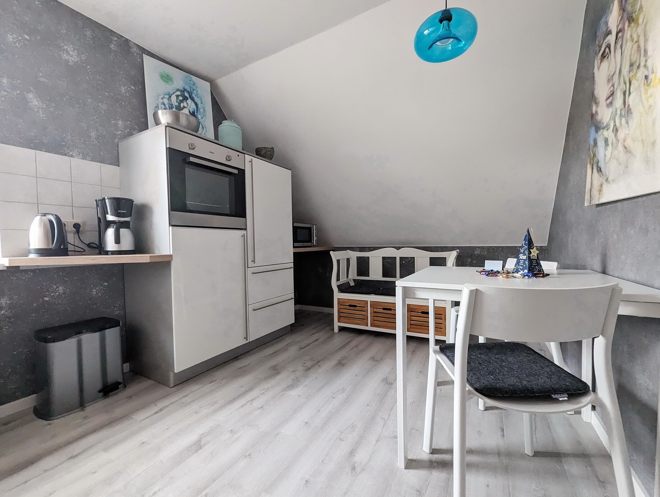 A compact kitchen with a microwave, kettle, and coffee maker on a white countertop. There is a white dining table with a chair and a small blue hanging light fixture above it.