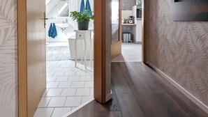 Un interno moderno con un corridoio che conduce a una zona pranzo, caratterizzato da un pavimento in legno, una parete bianca con un motivo a foglia e un soffitto con un lampadario.