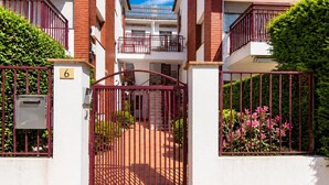 Edifício residencial com portão, placa com o número 6, escadaria que leva a uma varanda e um pequeno jardim com flores cor-de-rosa.