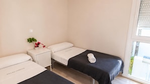 Quarto de hotel com duas camas, uma pequena mesa de cabeceira com uma planta e uma janela com persianas.