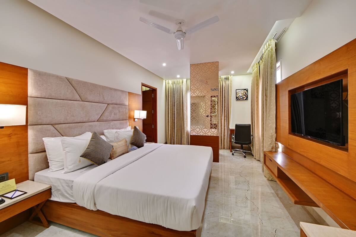 A modern hotel room with a large bed, wooden headboard, a flat-screen TV, a desk with a lamp, and a ceiling fan.