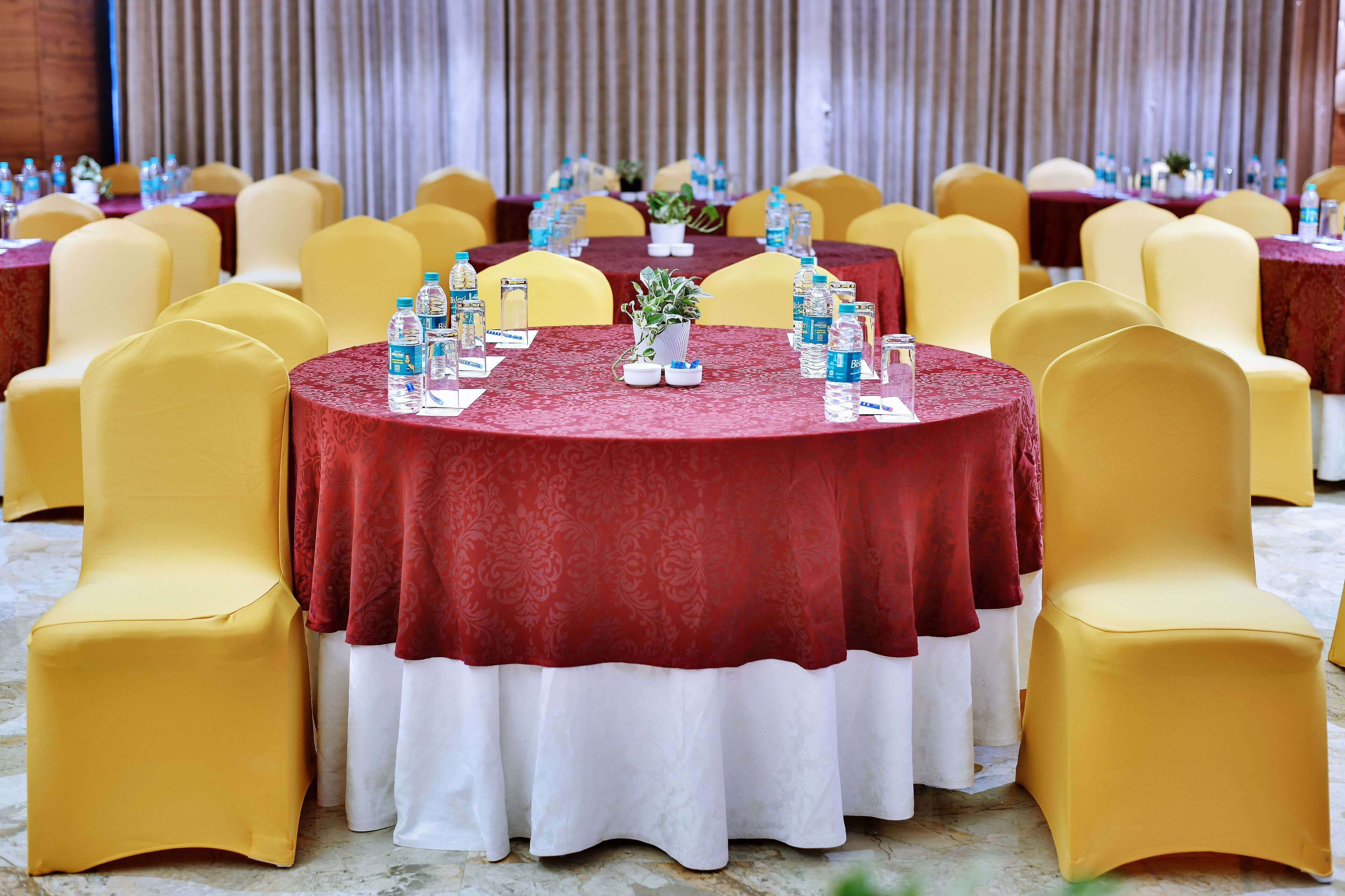 A banquet hall with round tables covered in red tablecloths, yellow chairs, and water bottles on each table.