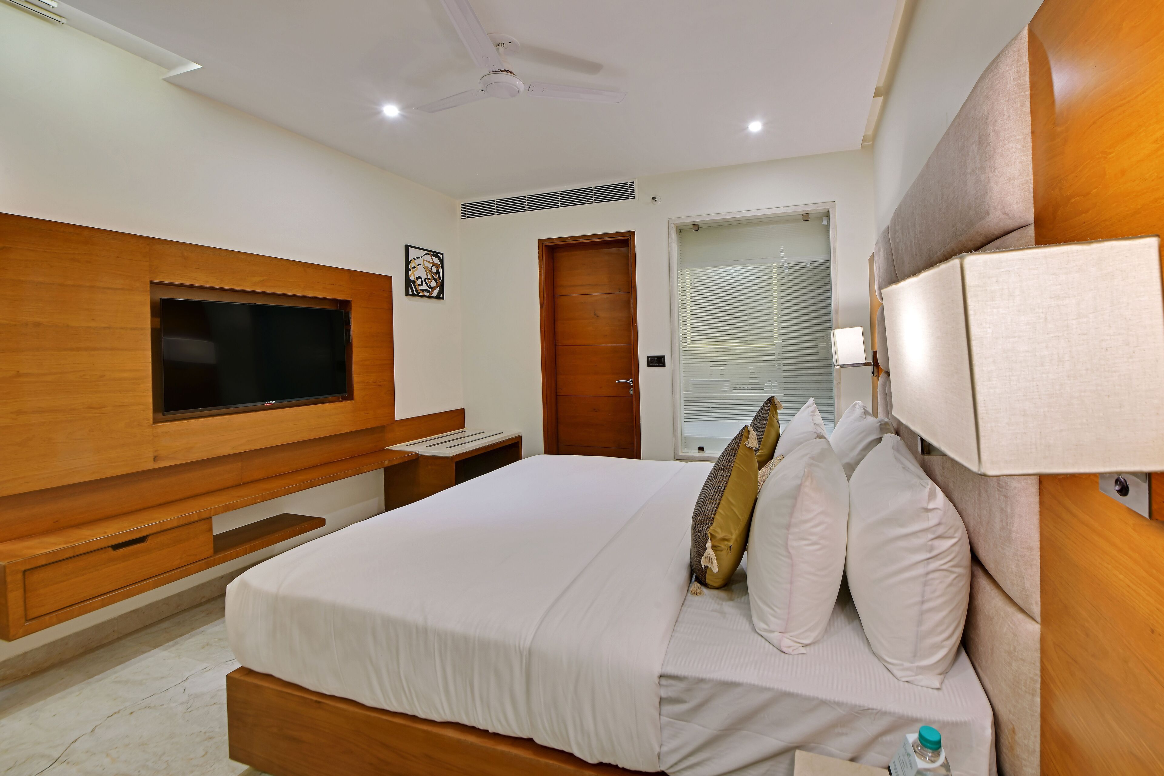 A modern hotel room with a large bed, wooden headboard, built-in TV, bedside table, and a window with blinds.