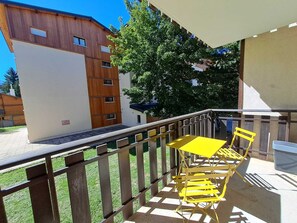 Ein Balkon mit gelben Stühlen und einem Tisch, der auf ein Gebäude mit Holzfassade und eine Rasenfläche blickt.