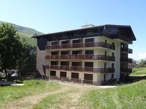A multi-story building with balconies, surrounded by greenery and hills.