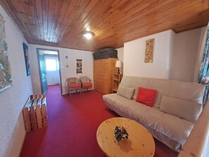 Een kamer met een houten plafond, een beige bank met rode kussens, een houten salontafel met een bloemstuk, en een houten garderobe.