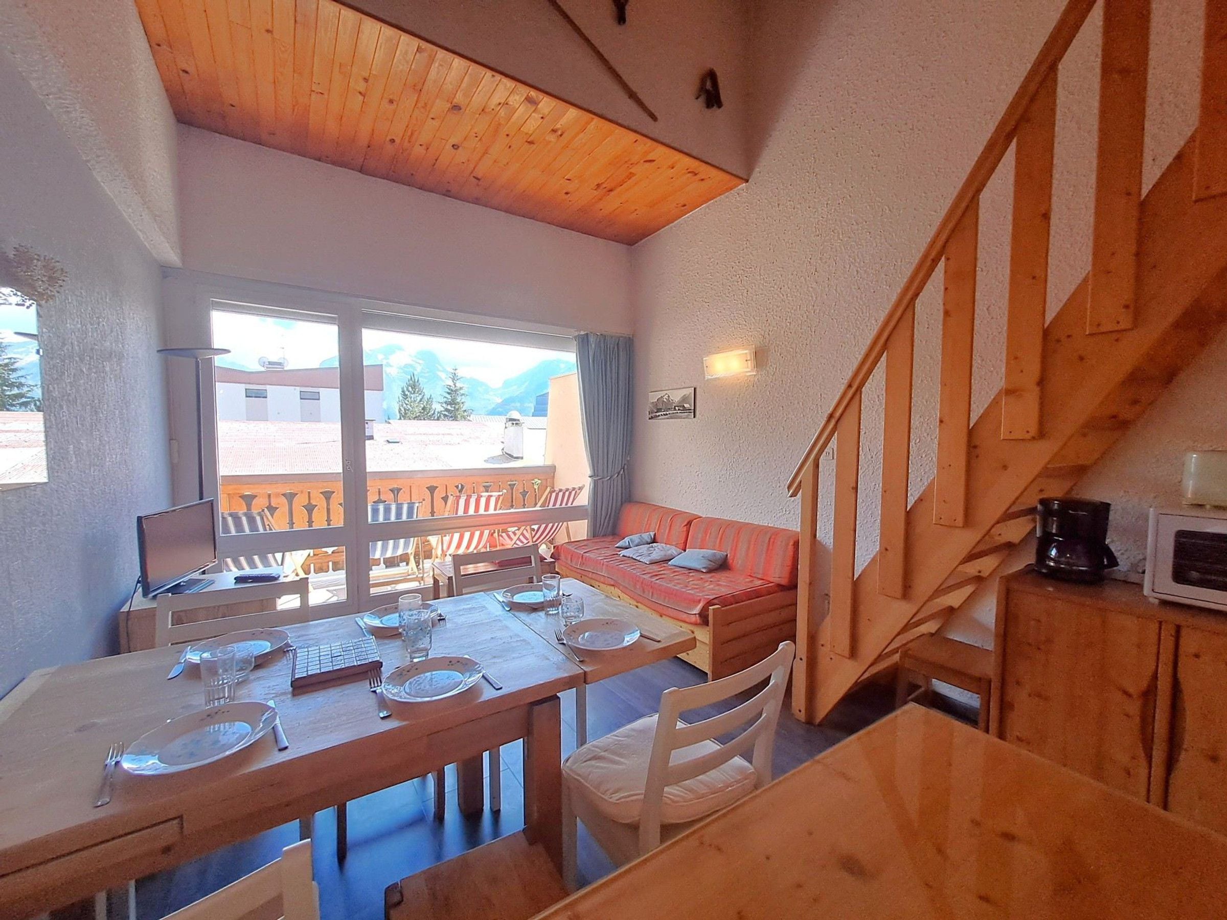 A dining area with a wooden table set for two, a red sofa, and a staircase with wooden railings.
