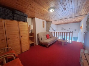 Ein gemütliches Zimmer mit rotem Teppich, Holzmöbeln, einer Couch mit rotem Kissen und einem Balkon mit Geländer.