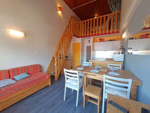 Een knus woongedeelte met een houten trap, een eethoek met een tafel en stoelen, en een kitchenette met kasten en apparatuur.
