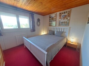Ein Schlafzimmer mit einem Einzelbett, einem Fenster mit Vorhängen, einem Heizkörper und einem an der Wand befestigten Regal mit Bildern.