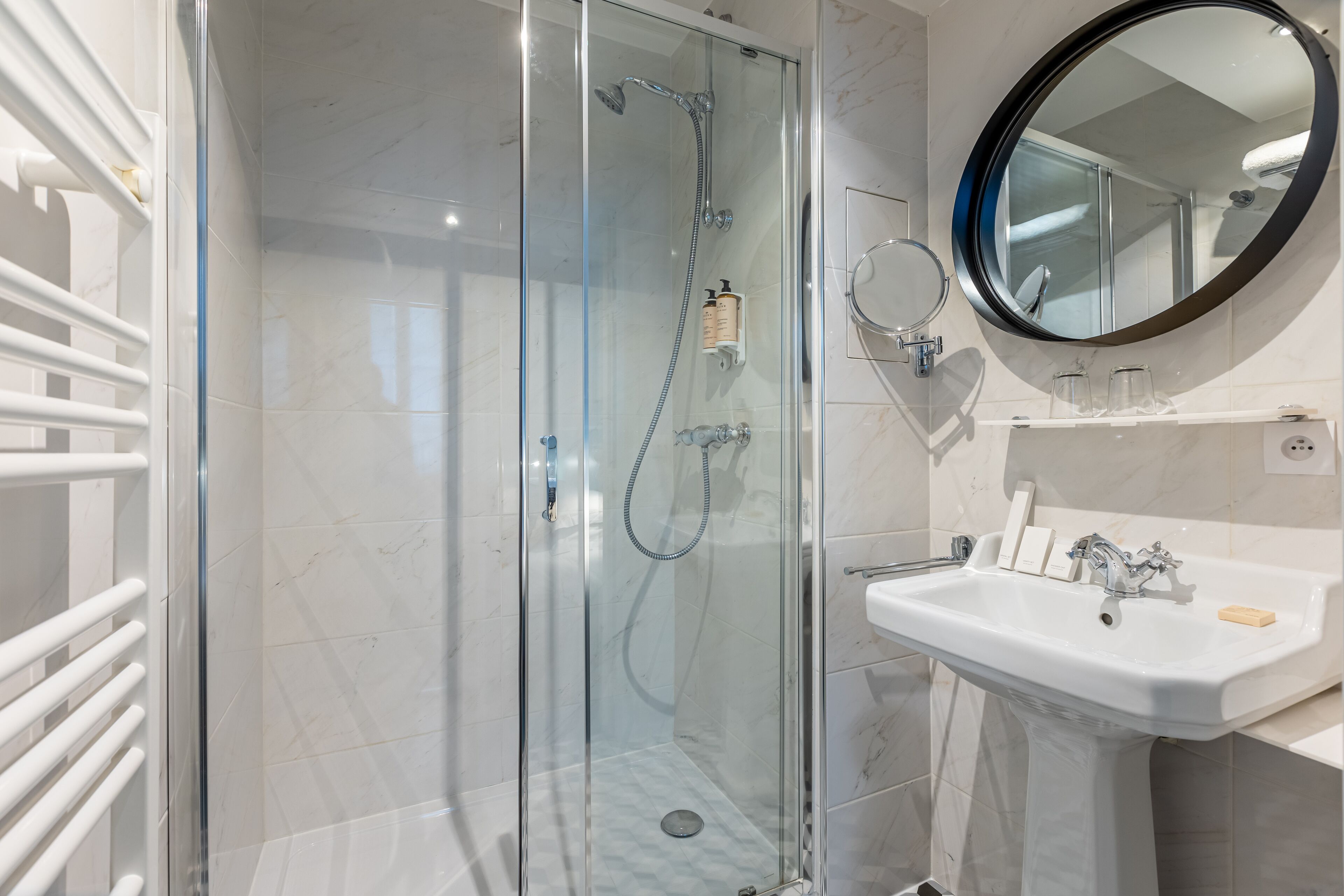 A modern bathroom with a glass-enclosed shower, a white pedestal sink, and a round mirror.