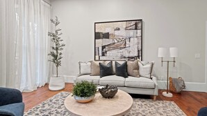 Ein Wohnzimmer mit einem hellen Sofa, einem runden Couchtisch mit Dekoration, einem großen abstrakten Gemälde an der Wand und einer Stehlampe.