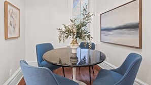 Un coin repas comprenant une table ronde en verre, quatre chaises bleues, un vase de fleurs et deux œuvres d’art encadrées accrochées aux murs.