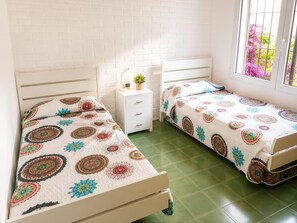Een kamer met twee eenpersoonsbedden, een wit nachtkastje met een lamp, een kleine potplant en een raam met uitzicht op roze bloemen.