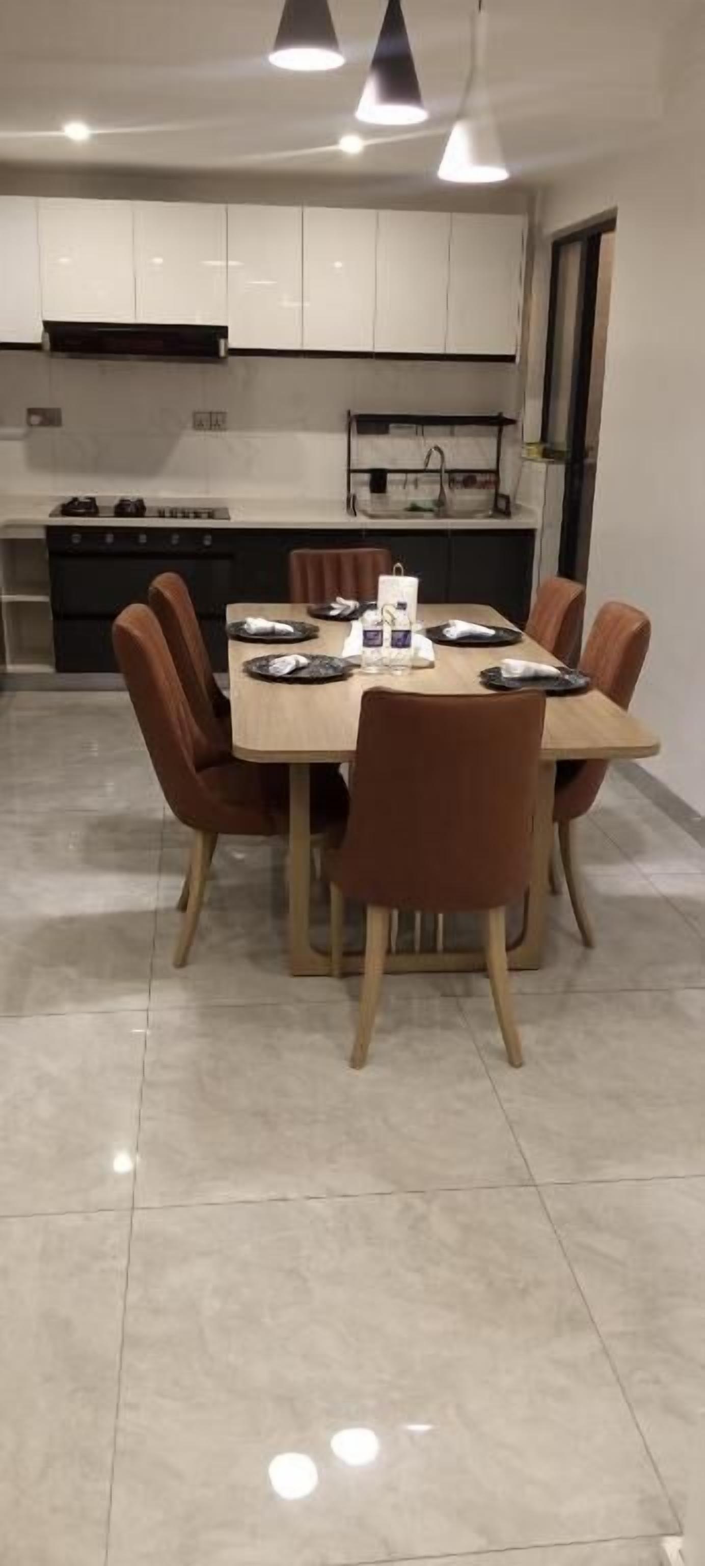 Een moderne eetruimte met een houten tafel en bruine stoelen, een keuken op de achtergrond en een betegelde vloer.
