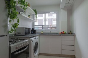 Et kompakt køkken med gasblus, mikroovn og vaskemaskine. Der er et vindue med udsigt til bygninger.