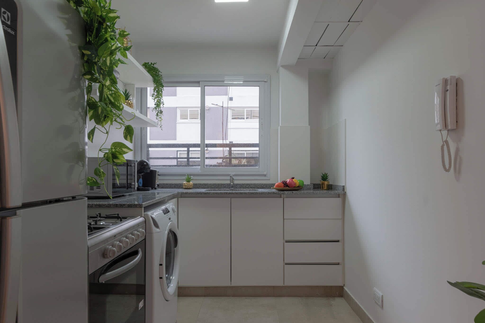 Une cuisine compacte équipée d’un four encastré, d’un lave-linge et d’une fenêtre donnant sur des immeubles.