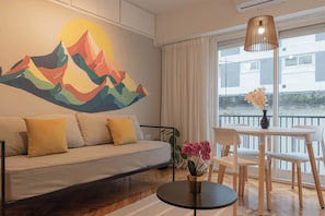 A modern living room with a sofa, a dining table, and chairs. There is a large window with curtains, a wall-mounted lamp, and a colorful mountain mural.