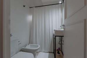 Pieni, valkoinen kylpyhuone, jossa on suihkuverho, peilillä varustettu pesuallas ja wc.