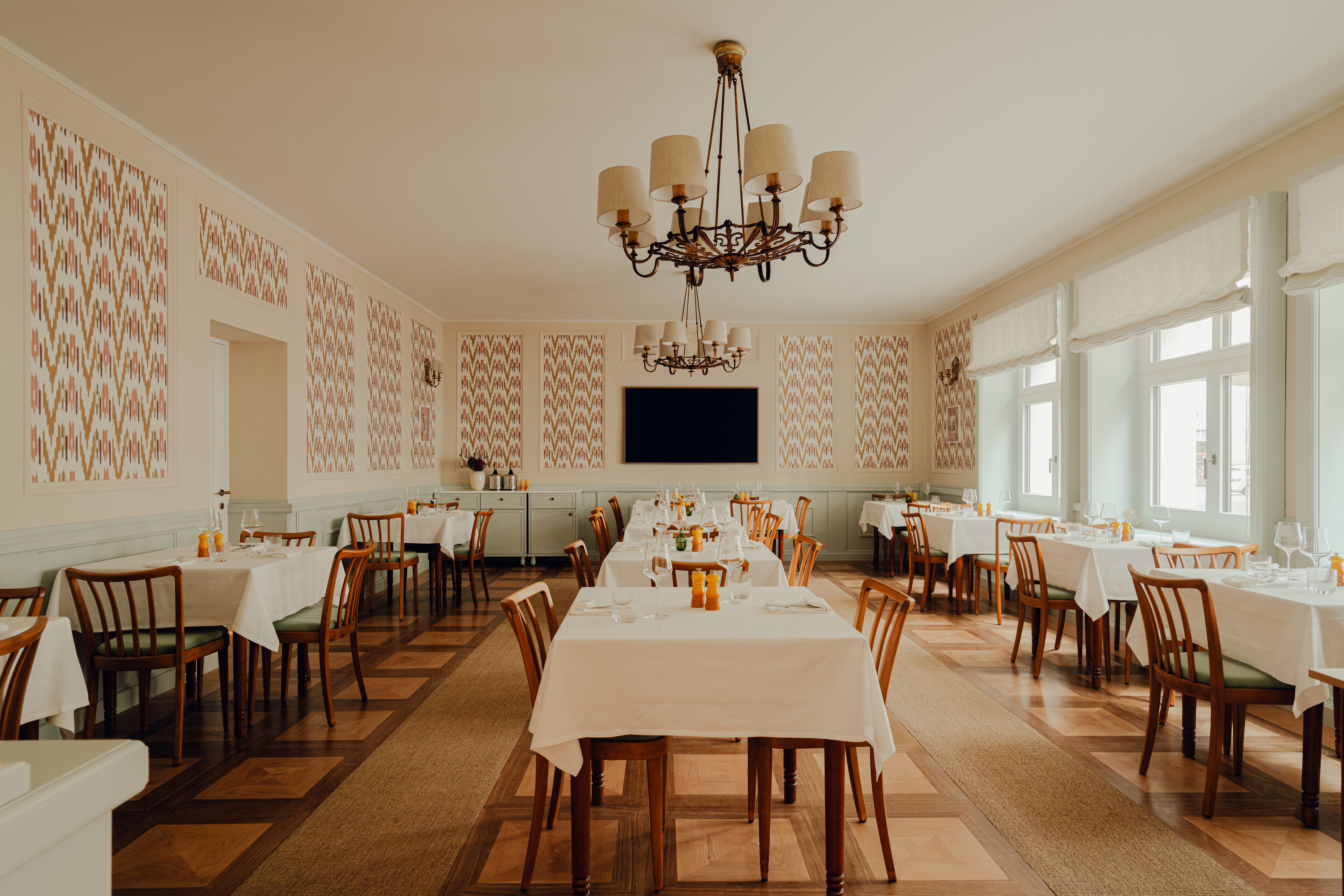 A spacious dining area with multiple tables and chairs, a chandelier, and patterned wallpaper.
