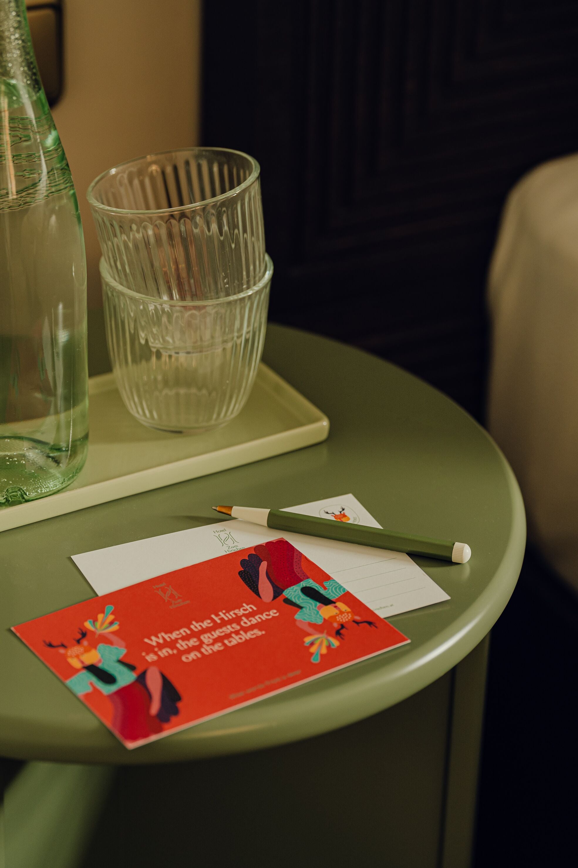 A green table with a glass bottle, a glass, a pencil, and a red card with a dance illustration.