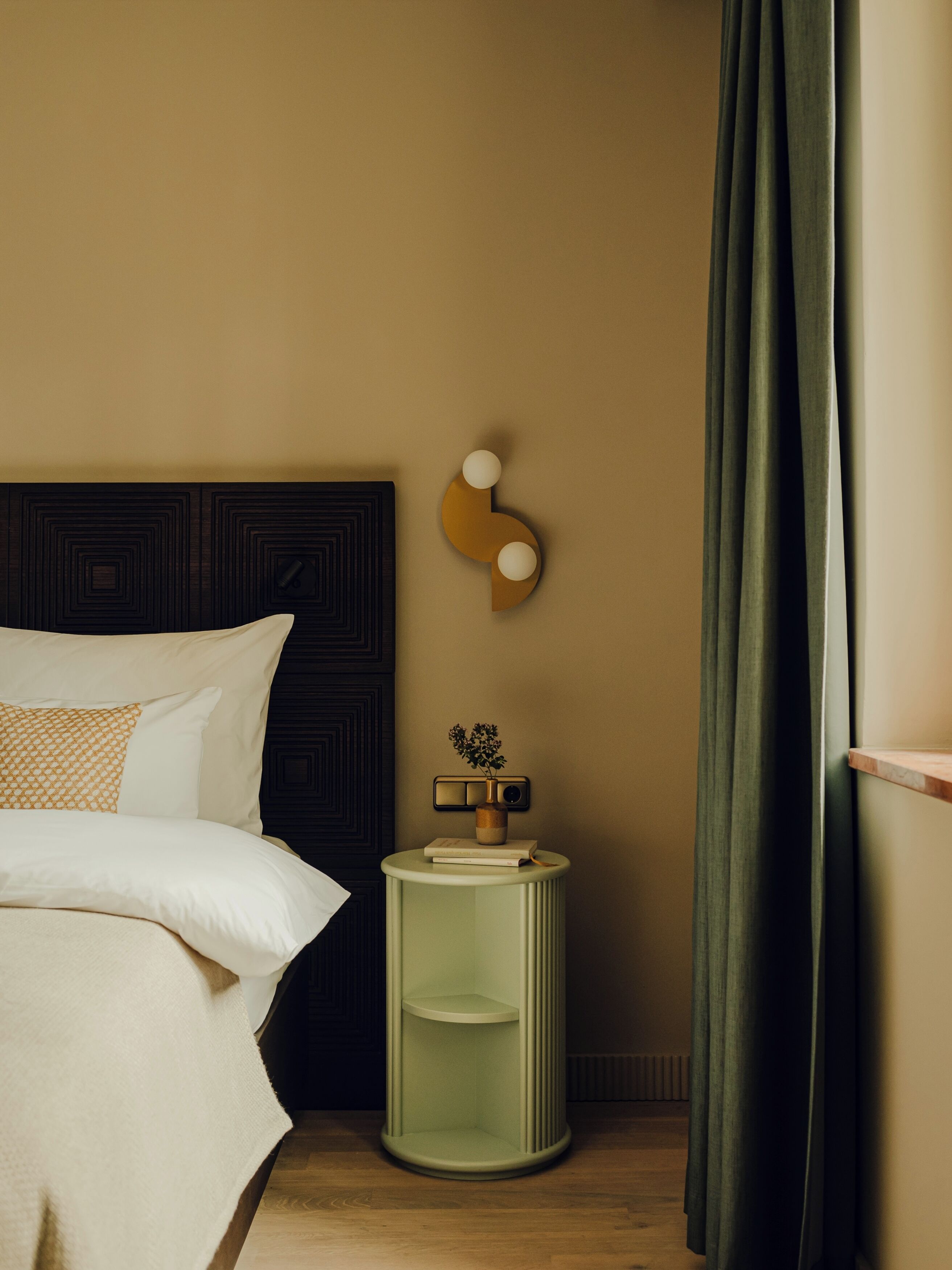 A neatly made bed with a patterned pillow, a bedside table with a vase and a book, and a wall-mounted light fixture.