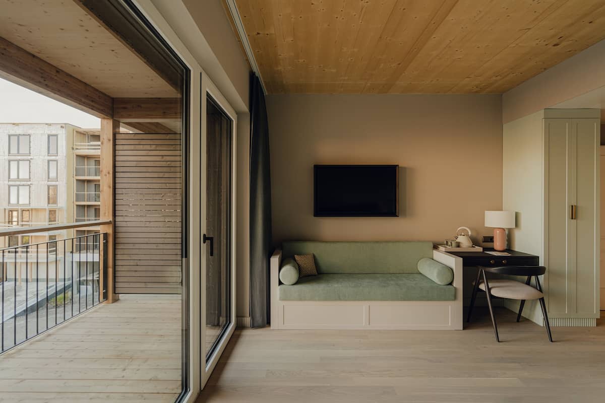 Une chambre moderne avec un plafond en bois, un canapé vert, un bureau avec une chaise et une grande fenêtre donnant sur un immeuble.