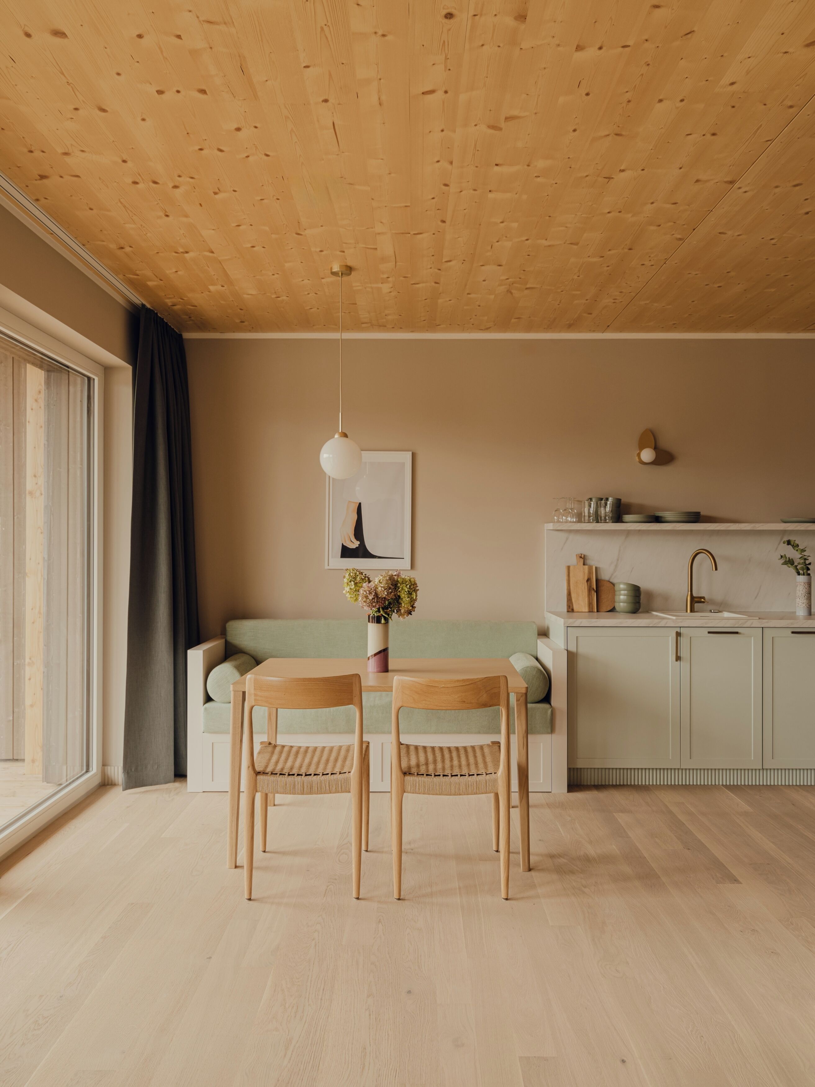 Une cuisine moderne avec des meubles en bois, une table à manger avec des chaises, un luminaire suspendu et une fenêtre avec des rideaux.