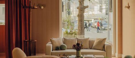 A cozy living room with a sofa, a coffee table with books and a vase, and a view of a tree and street outside.