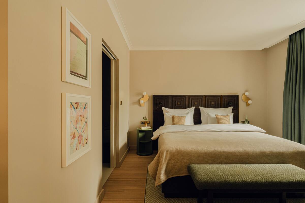Une chambre d’hôtel avec un grand lit, une table de chevet, un banc et deux lampes fixées au mur.