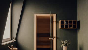 Una habitación con techo inclinado, puerta de madera, un estante pequeño con una botella, un banco, una planta en maceta y un estante fijado a la pared.
