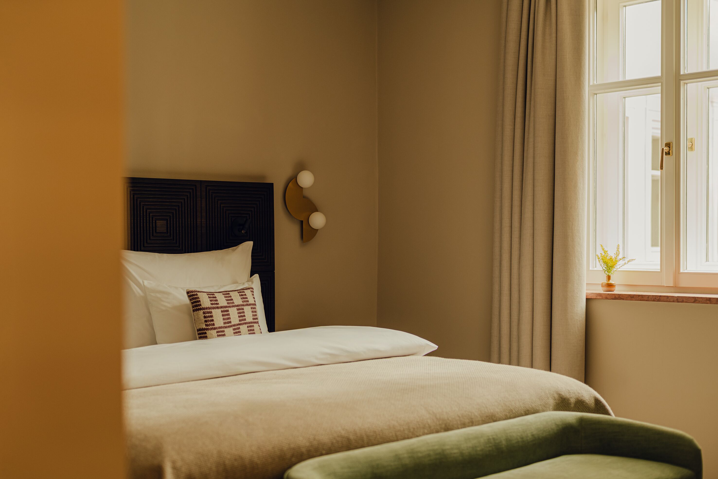 Una cama bien hecha con cabecera, mesita de noche y una ventana con cortinas.