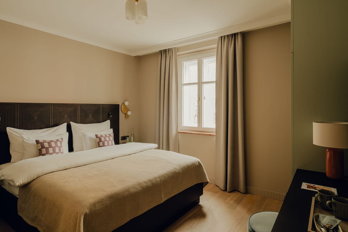 Une chambre d’hôtel avec un grand lit, un bureau avec une lampe, une fenêtre avec des rideaux et une horloge fixée au mur.