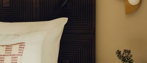 A neatly made bed with pillows, a dark patterned headboard, a bedside table with a vase and a book, and a wall-mounted light fixture.