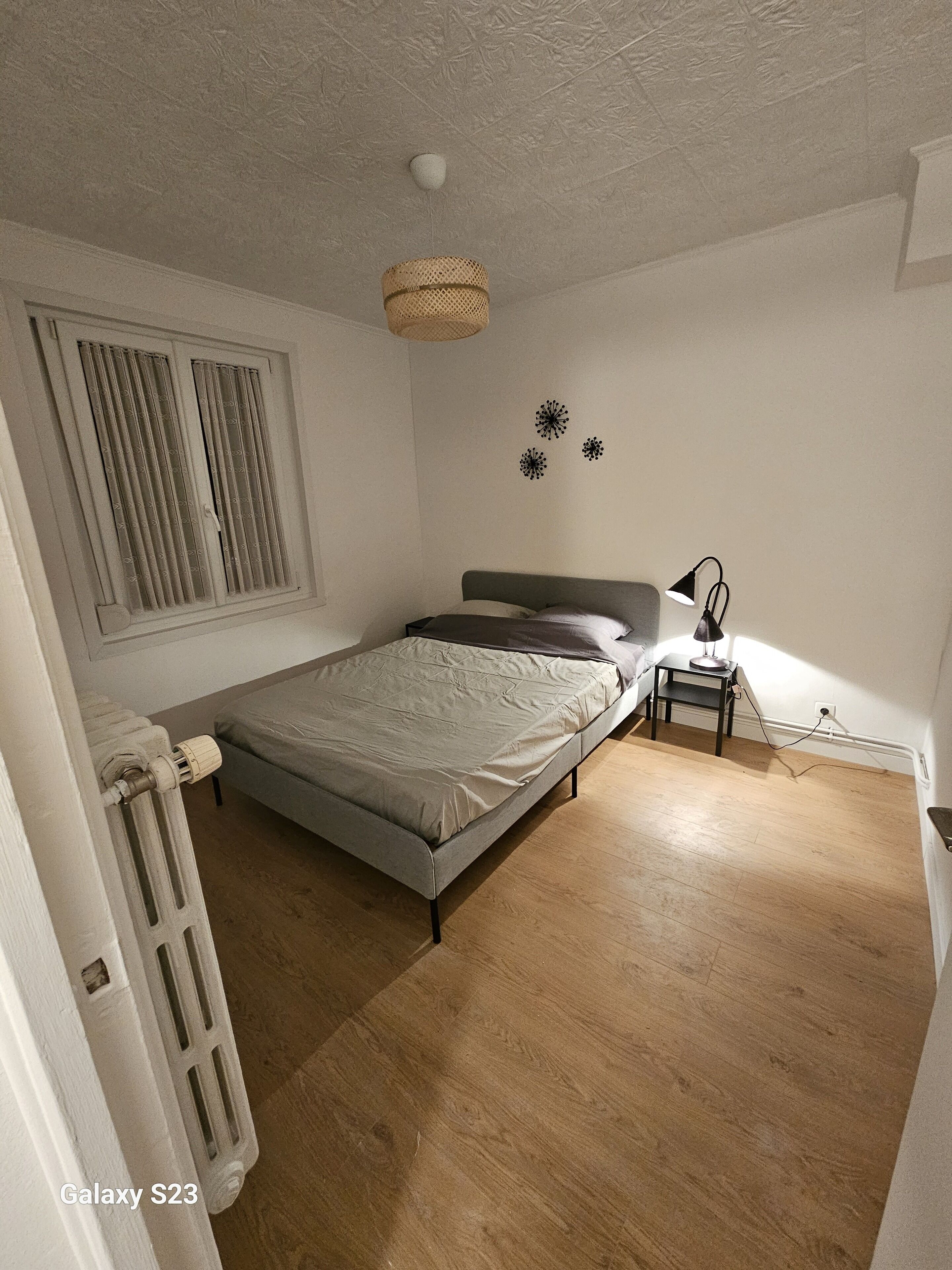 A bedroom with a bed, a nightstand, a lamp, a radiator, and a window with curtains.