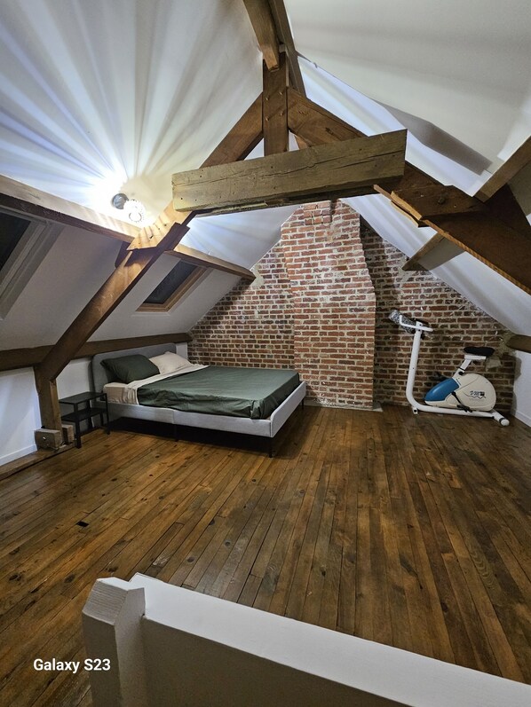 Une chambre à coucher aménagée dans un espace de type « loft », avec un plafond en pente, des murs en briques apparentes, un plancher en bois, un lit et un vélo d’exercice.