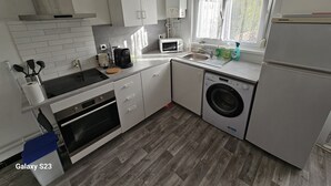 Een compacte keuken met witte kasten, een inbouwoven, een magnetron, een wasmachine en een raam met gordijnen.