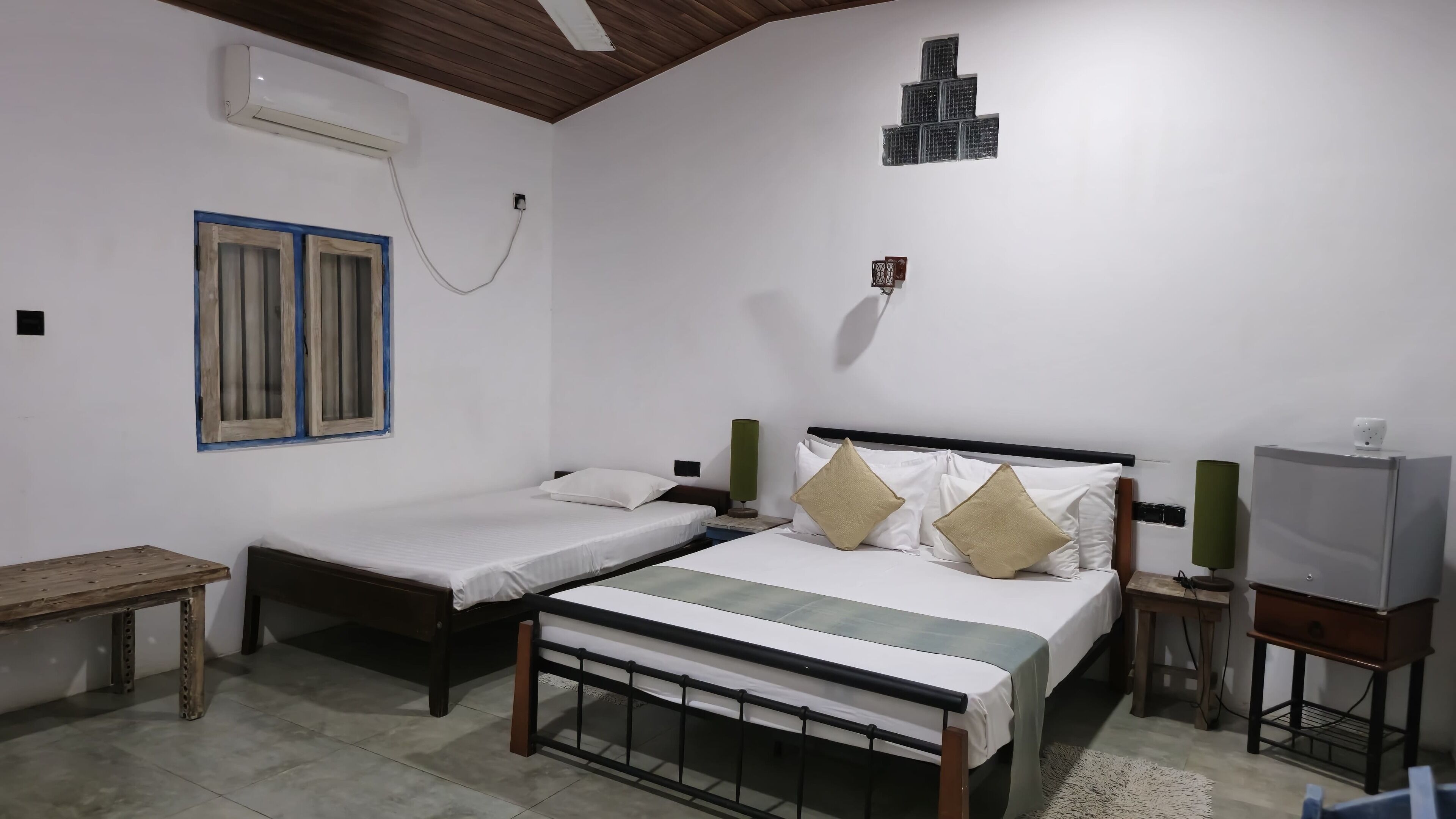 Un dormitorio con dos camas individuales, techo de madera, aire acondicionado de pared, una ventana con persianas y una mesita.