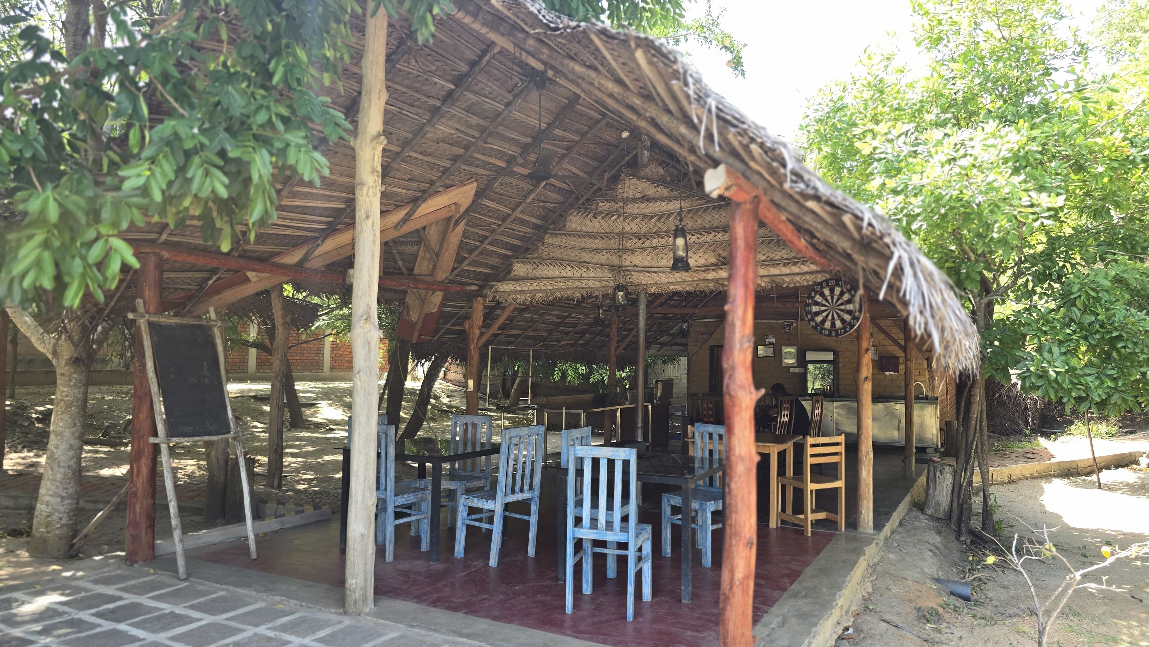 Un espacio de estar al aire libre rústico con mesas y sillas de madera, techo de paja y un dardo.