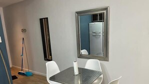 A dining area with a glass-top table, four white chairs, a large mirror, and a broom in the corner. - Muy Bien Ubicado y Comodo (ABN)